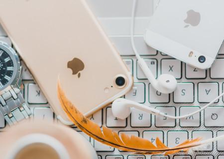 A pink iPhone, a white iPhone, a gold feather, and earbuds lying on top of a keyboard.